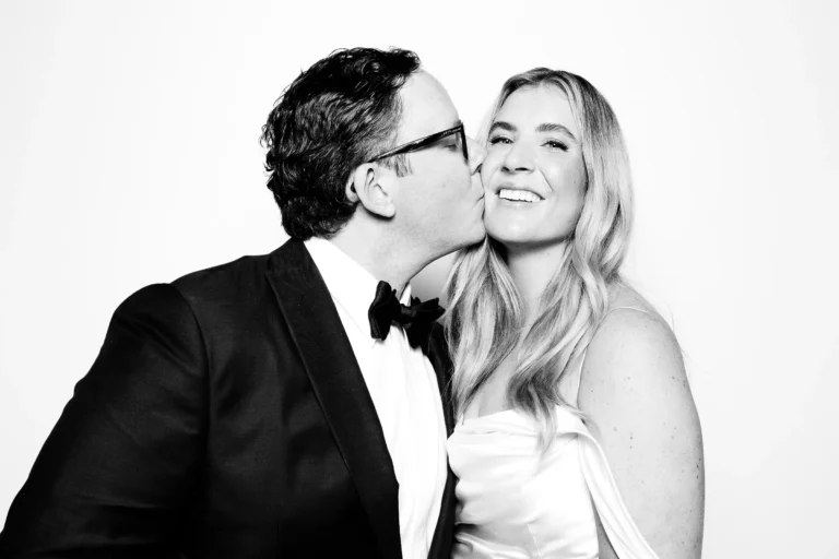 Wedding photo booth rental 2 Guest in a tuxedo kisses a smiling partner in a white satin dress on the cheek in black and white.