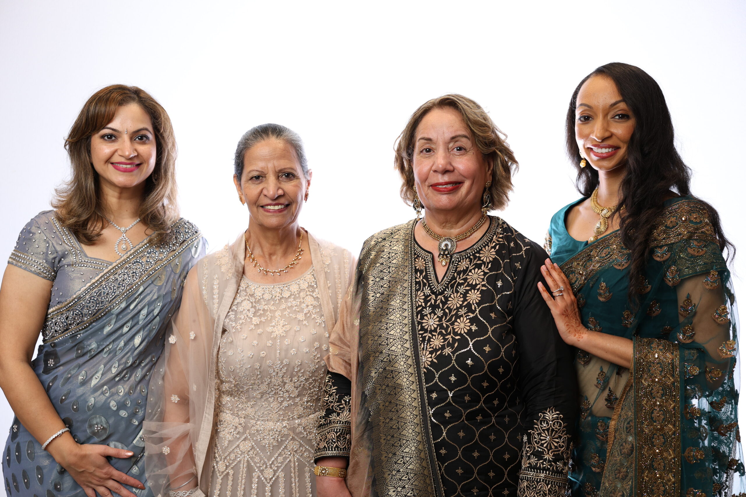 Four women standing side by side against a white background, smiling at the camera and wearing elegant traditional South Asian formal attire with intricate embroidery, jewelry, and draped fabrics, posed for a studio-style portrait.