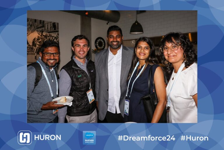 Five smiling guests pose with drinks in a blue branded event photo frame.