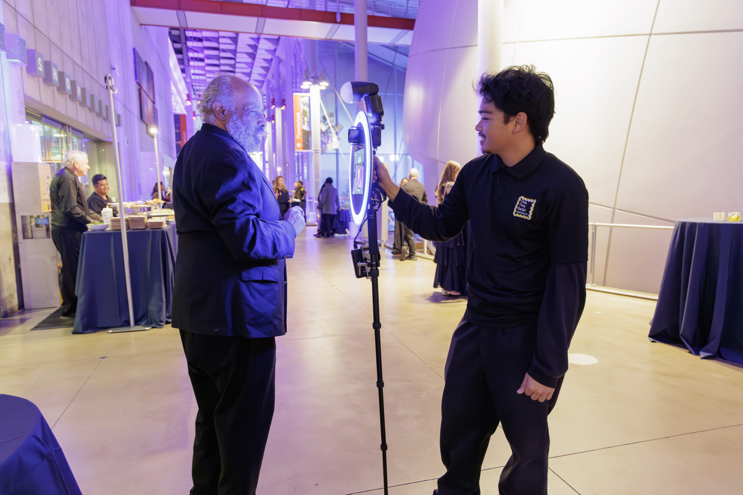 A One, Two, Smile! Photobooth attendant assists a guest at a ring-light photo station in a purple-lit event hall.