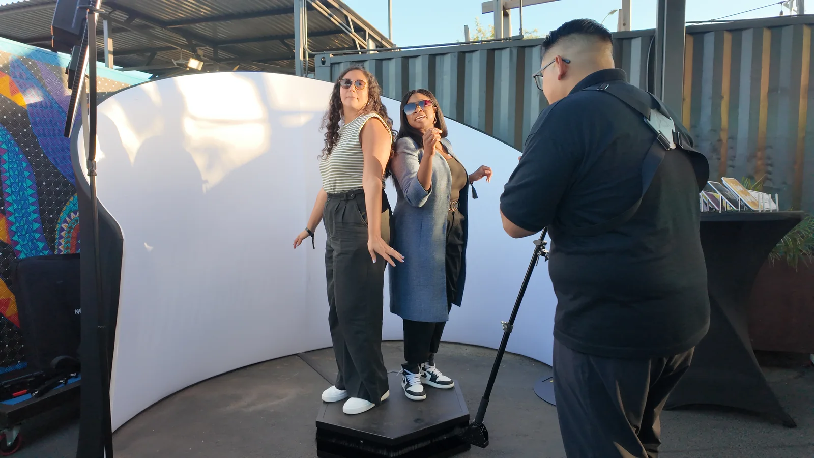 Two guests stand on an outdoor 360 photo booth platform in front of a curved white backdrop.