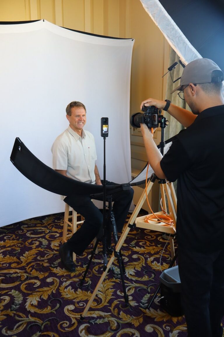 Photographer photographs a seated guest with a curved reflector and white backdrop.